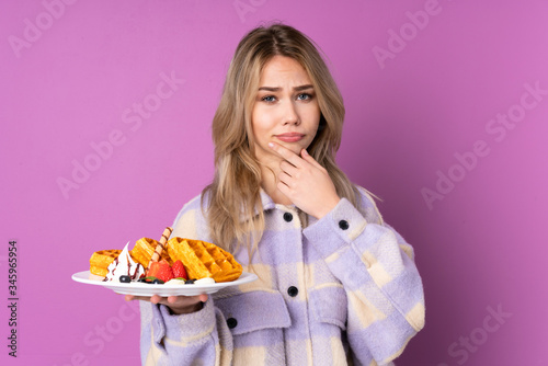 Teenager Russian girl holding waffles isolated on purple background thinking