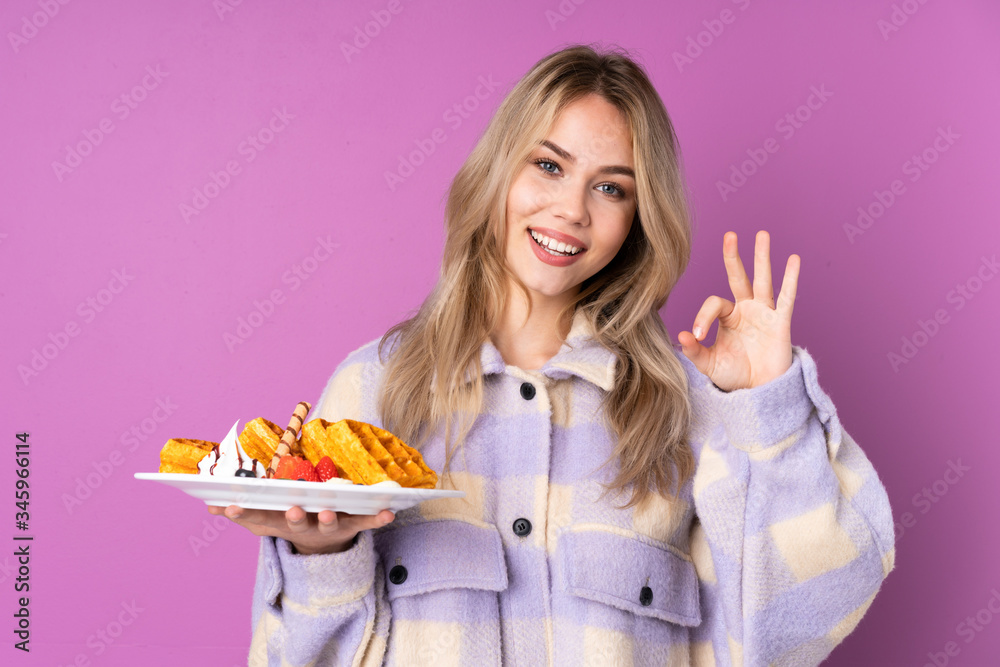 Teenager Russian girl holding waffles isolated on purple background showing ok sign with fingers