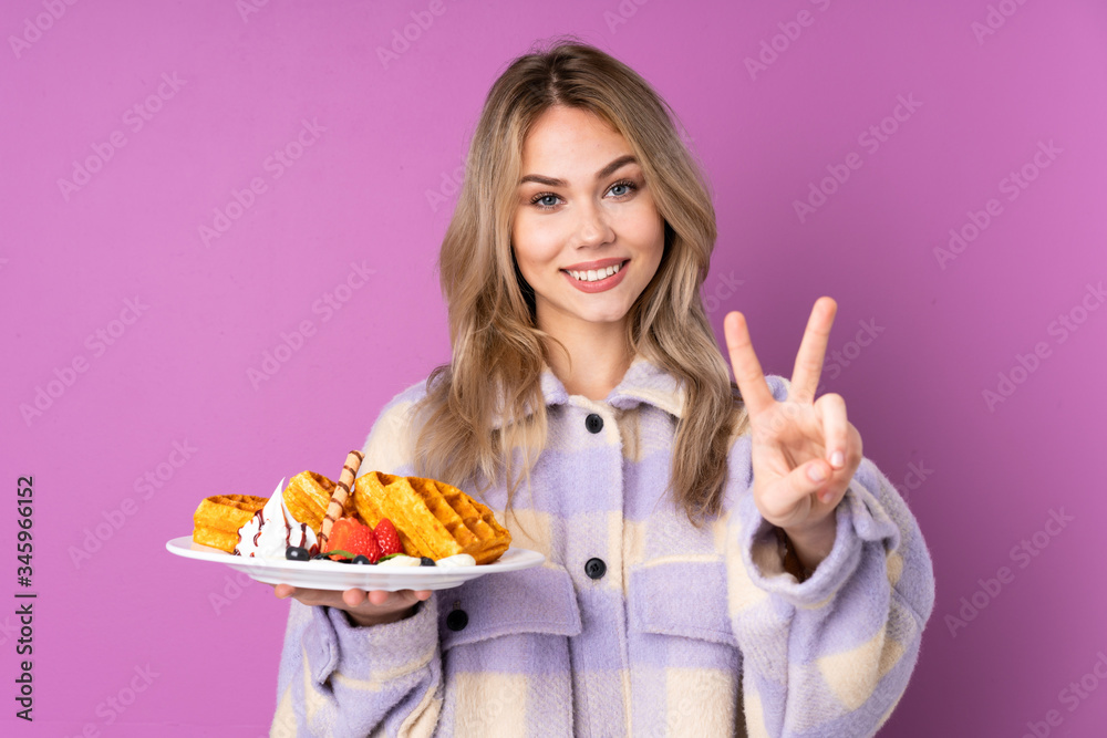 Teenager Russian girl holding waffles isolated on purple background smiling and showing victory sign Teenager Russian girl holding waffles isolated on purple background smiling and showing victory sign
