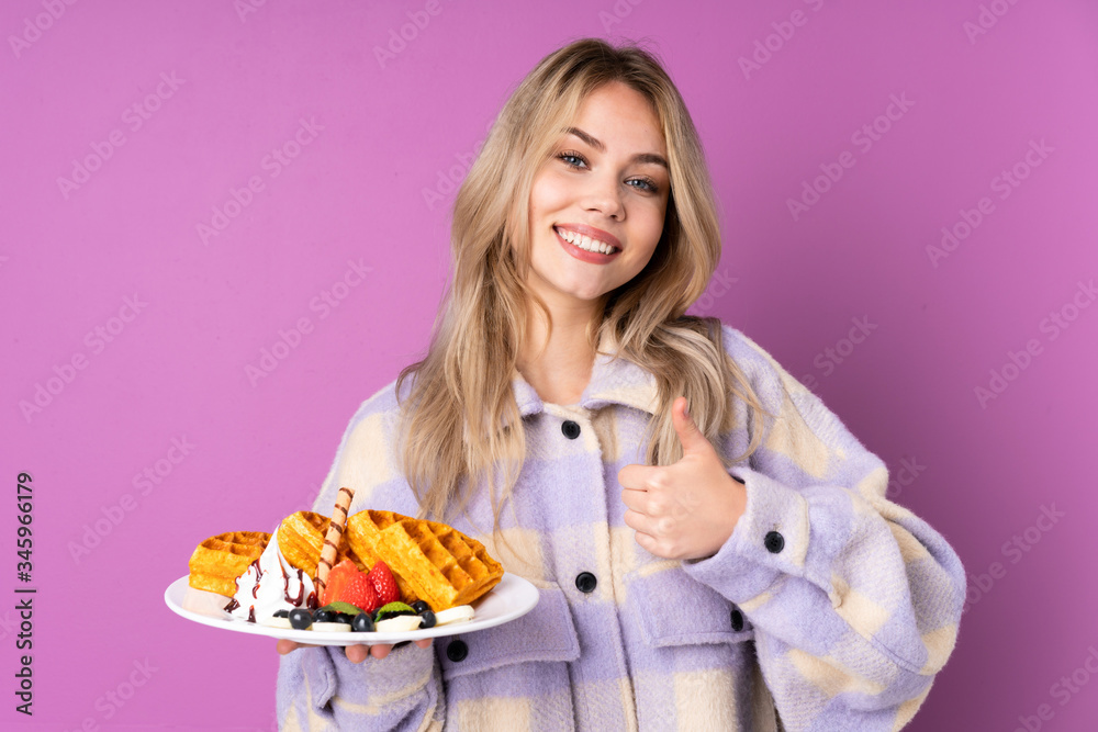 Teenager Russian girl holding waffles isolated on purple background giving a thumbs up gesture