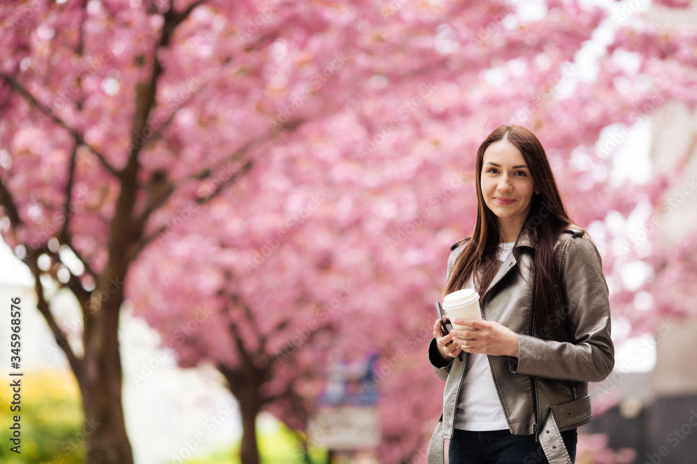 Girl in stylish outfit smiling and holding coffee on sakura background. Portrait of woman in jacket with cup of tea Girl in stylish outfit smiling and holding coffee on sakura background. Portrait of woman in jacket with cup of tea