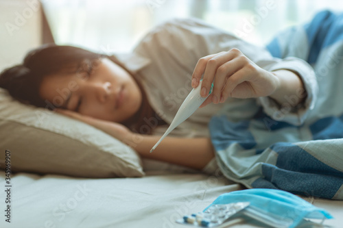 Obraz na płótnie Asian teen infected with Covid-19 flu sick lying in bed due to a Corona virus pandemic, anxiously measuring check body temperature with digital thermometer