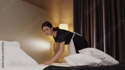 young, beautiful maid in uniform and apron making bed in hotel room