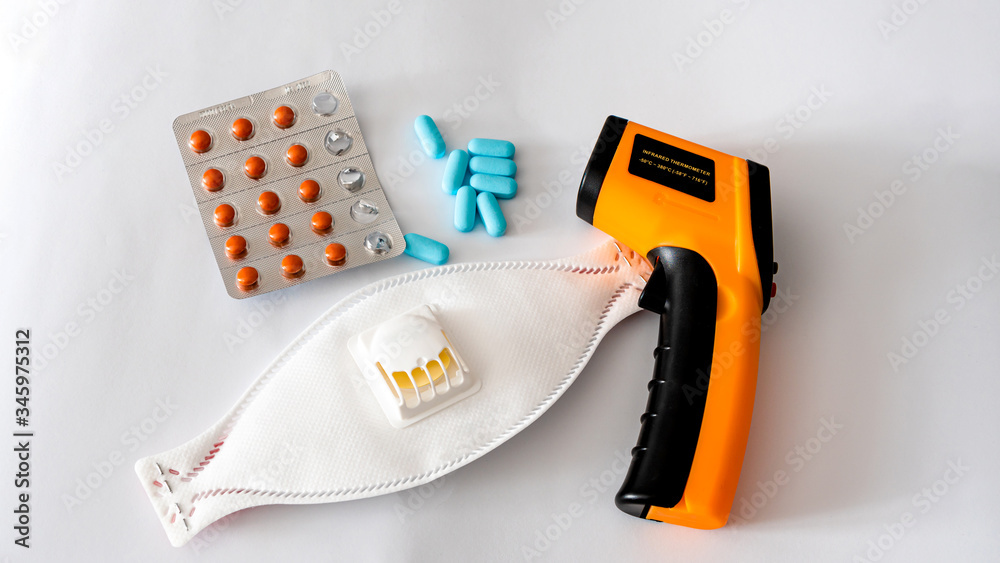 Remote electronic thermometer on a white background next to pills and a respirator mask.