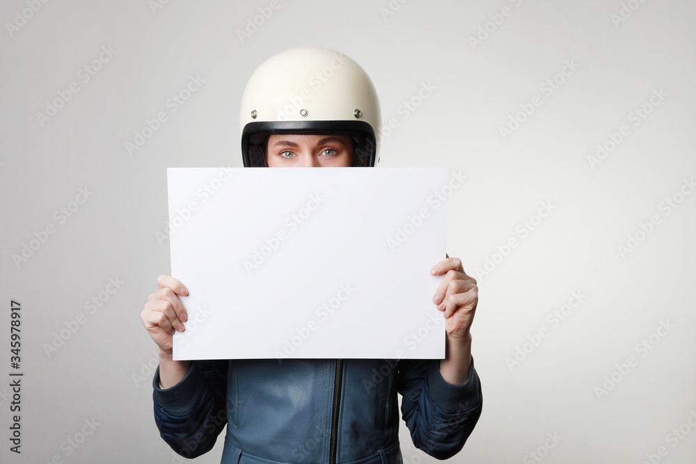 Shot of young biker woman, keeping a white banner for advertising, wears leather jacket, over studio background.