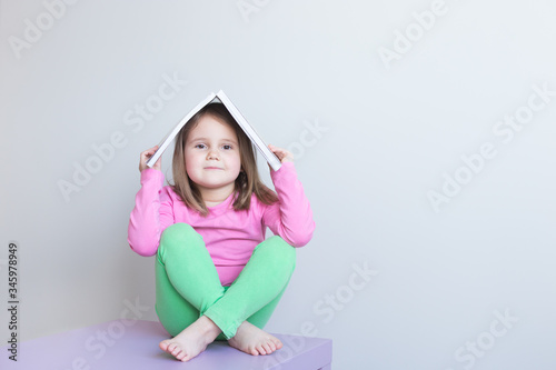 Portrait of a white girl 4 years old. Pink and green clothes. She holds a book in the shape of a house on her head. Gray background Selective focus. Copyspace.