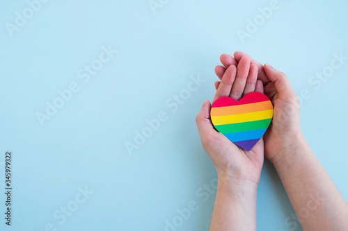 Womans hands hold heart in the colors of rainbow on light blue background. LGBT concept, Pride month. Top view, copy space