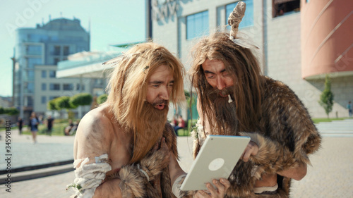 Ancient wild homo sapiens rejoicing with new toy technology digital tablet outdoors. Primitive timetravelers of hunter-gatherers tribe in modern cityscape.