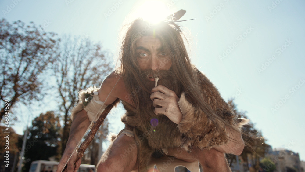 Aboriginal caveman tapping with stone-tipped spear on camera discovering new device chewing animal bone outside in future city. Savage in downtown. Aboriginal caveman tapping with stone-tipped spear on camera discovering new device chewing animal bone outside in future city. Savage in downtown.