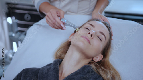 Head of beautiful woman receiving skin healh care facial treatment in spa salon. Close-up beautician rf-lifting customer face skin with modern hardware cosmetology.