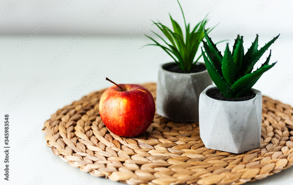 Cozy home interior decor: red apple and ornamental plants in pots on a wicker stand on a white table in the room. The quarantine concept of stay home. Cozy home interior decor: red apple and ornamental plants in pots on a wicker stand on a white table in the room. The quarantine concept of stay home.