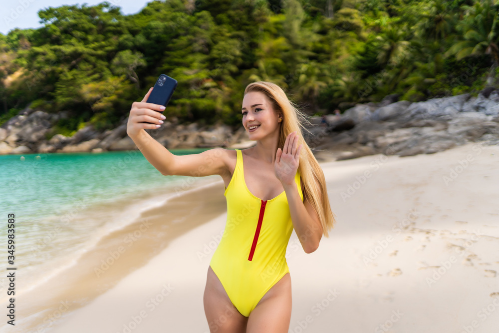 Portrait of a happy girl wearing yellow bikini laughing while watching streaming videos or make video call in smart phone on the beach with the ocean