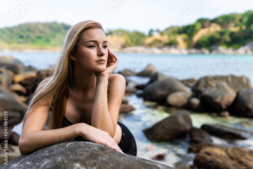 Young blonde woman in black bikini posing on a sand rocks near the sea