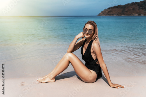 Young woman in black bikini sits on sandy beach poses against beautiful ocean view, enjoys summer time.