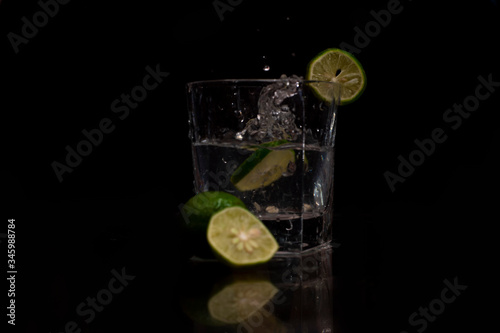 Water splashes out of the garnished glass as a green fresh lemon slice immerses into the water.