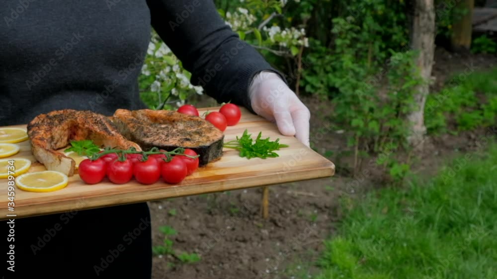 Waiter brings tray with baked salmon fish steak with tomatoes herbs lemon slice and spices. Grilled seafood with vegetables on wooden plate, green blooming trees background. Dining restaurant concept Waiter brings tray with baked salmon fish steak with tomatoes herbs lemon slice and spices. Grilled seafood with vegetables on wooden plate, green blooming trees background. Dining restaurant concept