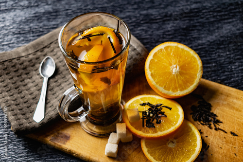 Citrus drink, tea with petals in a transparent glass mug with slices of sweet orange and refined sugar. Slicing fruit on a wooden Board with a brown napkin in a cozy dark background in a low key