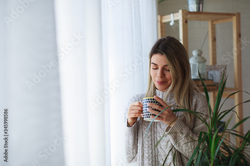 Young woman holding a cup of coffee standing by the window and looking out