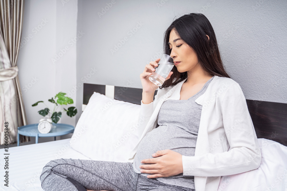 beautiful asian pregnant woman taking a medicine pill of vitamin and a glass of water sitting on white bed, relaxing and resting in a bed room as motherhood, wearing stretcher pants and white cardigan beautiful asian pregnant woman taking a medicine pill of vitamin and a glass of water sitting on white bed, relaxing and resting in a bed room as motherhood, wearing stretcher pants and white cardigan