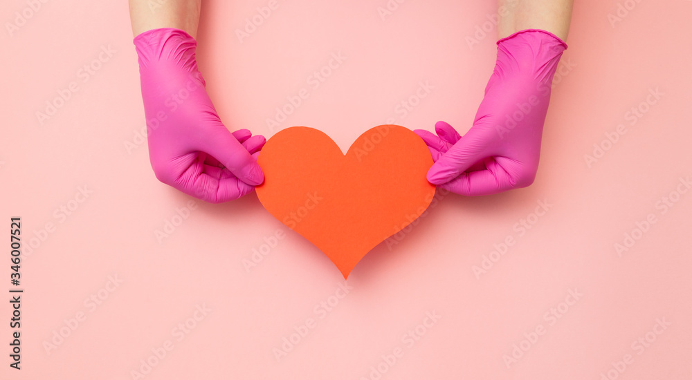 Sign of heart keeping female hands in medical red gloves on pink background. Banner with copy space for info about treatment assistance and thanks to the doctors. Sign of heart keeping female hands in medical red gloves on pink background. Banner with copy space for info about treatment assistance and thanks to the doctors.