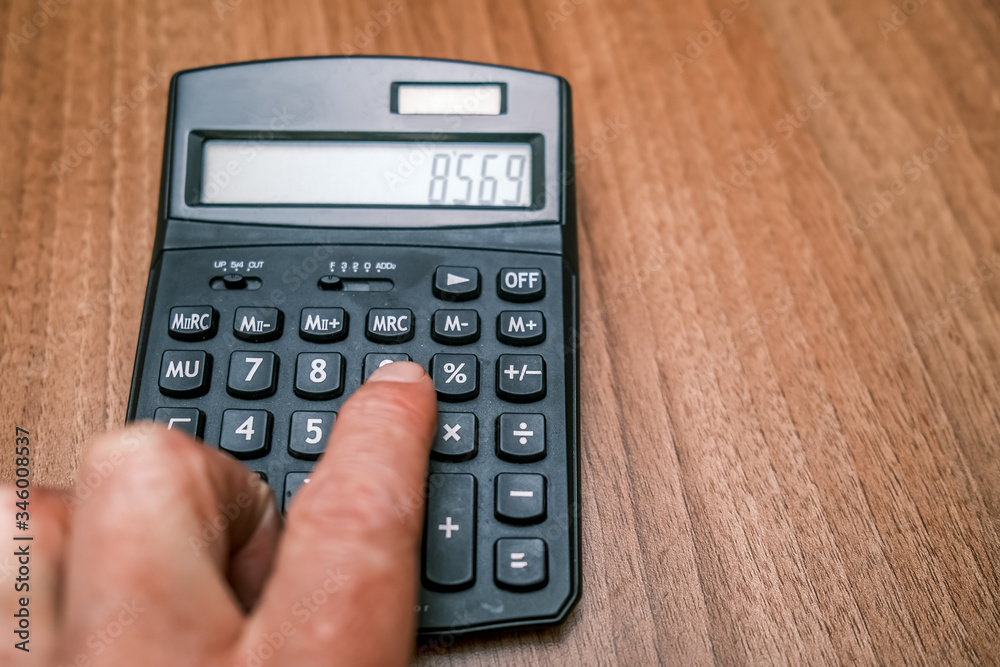 Close up of human hand using a black calculator laying on a wooden office desk Close up of human hand using a black calculator laying on a wooden office desk