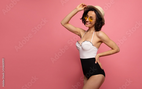 Portrait of a gorgeous trendy fit woman in swimwear while she enjoying rest with sunlight isolated over pink background. Travel, tourism, journey, trip, tour, summer concept