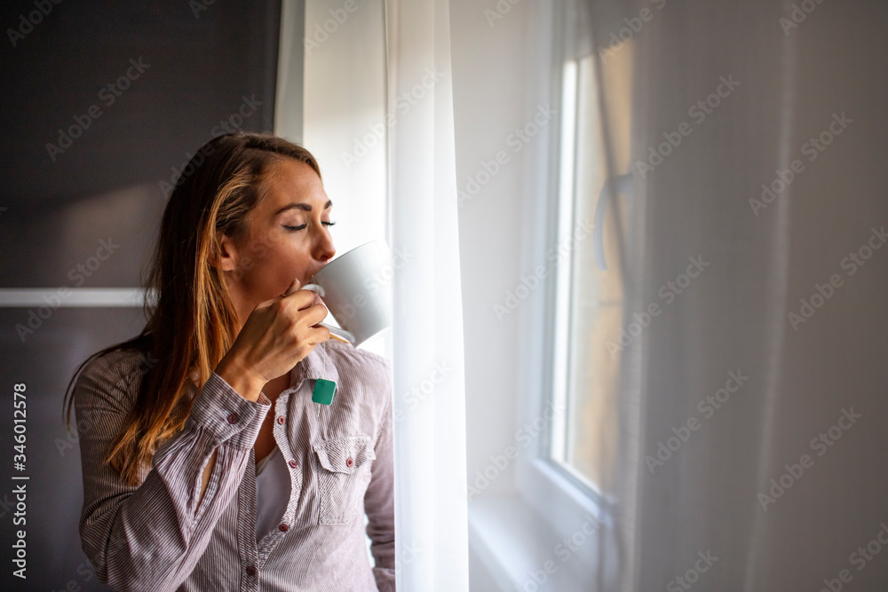 Beautiful woman standing near windows. Beautiful morning. Woman in the morning. Attractive woman is holding a cup with hot tea or coffee and looking at the window in her home Beautiful woman standing near windows. Beautiful morning. Woman in the morning. Attractive woman is holding a cup with hot tea or coffee and looking at the window in her home