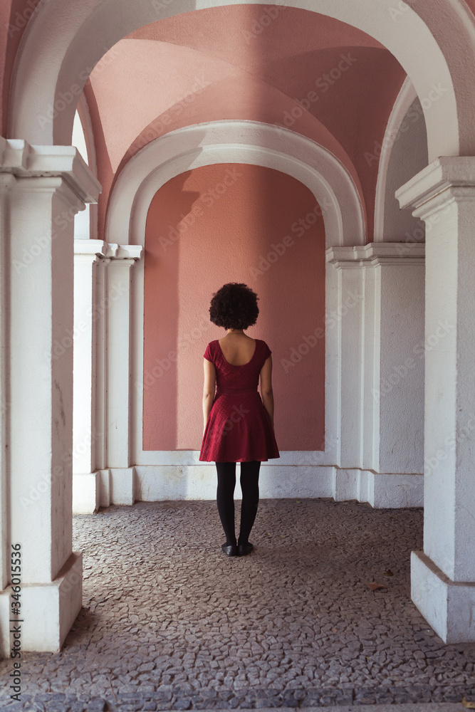 Black afroamerican young girl with curly hair standing backwards in front of a classic pink wall