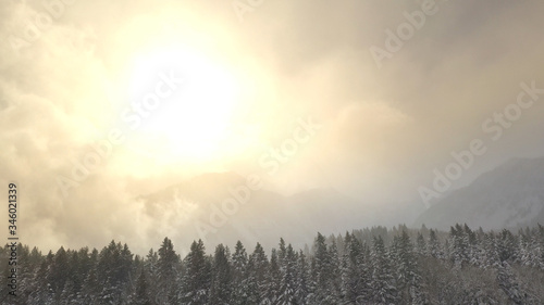 Flying up and over mountain forest covered in snow towards the sun as it lights up the mountain range below.