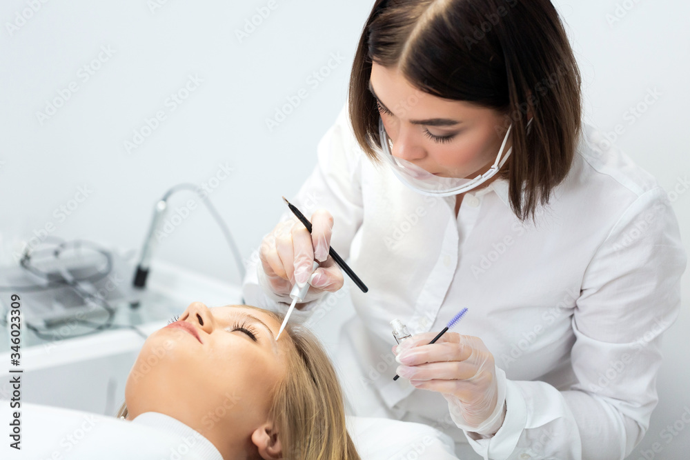 A young woman lies and master in a white uniform does eyebrow makeup in a beauty salon. The use of permanent makeup on eyebrows. The wizard works with eyebrows. Semi-permanent makeup. White background A young woman lies and master in a white uniform does eyebrow makeup in a beauty salon. The use of permanent makeup on eyebrows. The wizard works with eyebrows. Semi-permanent makeup. White background