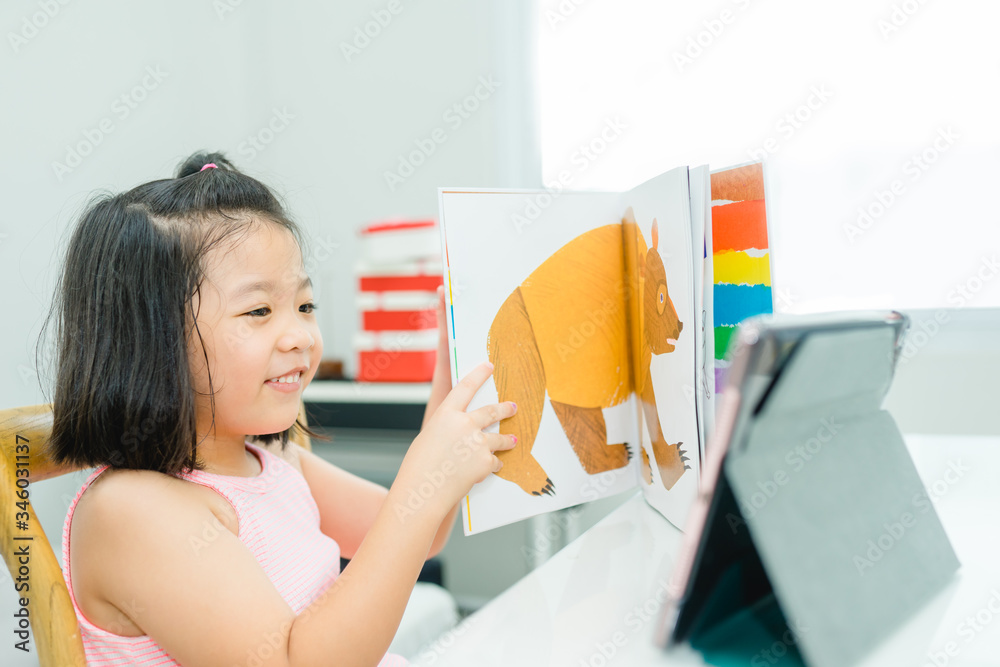 Φωτογραφία Asian girl student online learning class study online with video call to teacher, Happy young girl reading story book online and looking at tablet at home