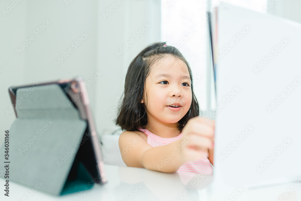Asian girl student online learning class study online with video call to teacher, Happy young girl reading story book online and looking at tablet at home.Covid-19 coronavirus.Social distancing. Asian girl student online learning class study online with video call to teacher, Happy young girl reading story book online and looking at tablet at home.Covid-19 coronavirus.Social distancing.