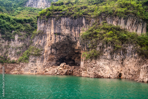 Wuchan, China - May 7, 2010: Dicui Gorge on Daning River. Deep cave in brown-black rocky cliff with green foliage on top. Emerald green water up front,