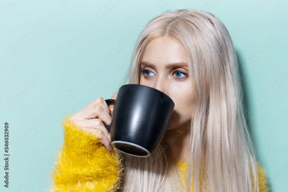 Φωτογραφία Portrait of young blonde girl with blue eyes drinking tea from black cup of background of aqua menthe color