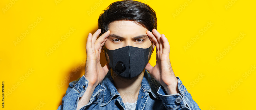 Studio portrait of young guy with hands on the head, wearing black respiratory face mask against coronavirus. Background of yellow color. Studio portrait of young guy with hands on the head, wearing black respiratory face mask against coronavirus. Background of yellow color.