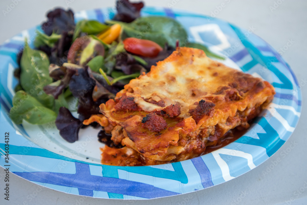 A meal of lasagna and a side green salad on a disposable paper plate