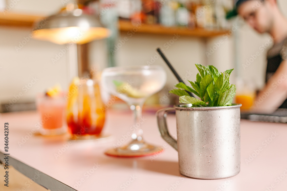 Selective focus on a metal cup with mint and three cocktail glasses prepared next to it on a bar