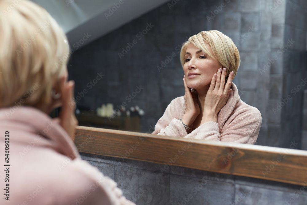 Skincare Routine. Mature Woman Touches Face Skin In Front Of The Mirror Reflection. Anti Aging Beauty Treatment Concept. Skincare Routine. Mature Woman Touches Face Skin In Front Of The Mirror Reflection. Anti Aging Beauty Treatment Concept.