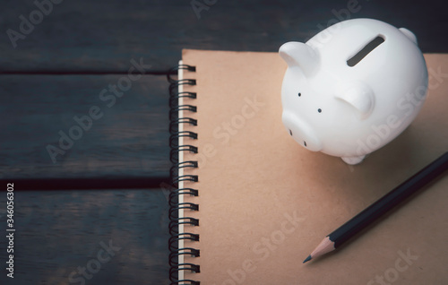 piggy bank on a paper notebook, which has a pencil for taking notes placed on a dark wooden table investment planning ideas, and money saving concept.