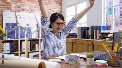 happiness female engineer celebrating success in office while got good news on laptop computer. woman architect employee cheerful laughing with raised arms in air. victory on construction project