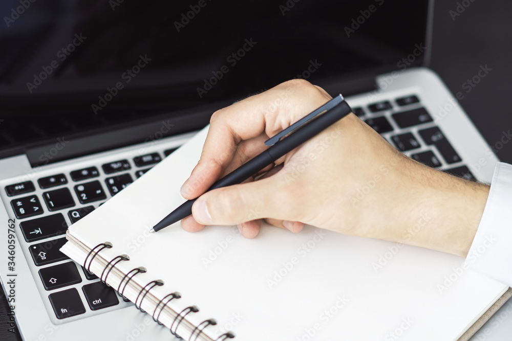 Man writes with a pen in notepad on laptop keyboard in a sunny office, business and education concept. Close up Man writes with a pen in notepad on laptop keyboard in a sunny office, business and education concept. Close up