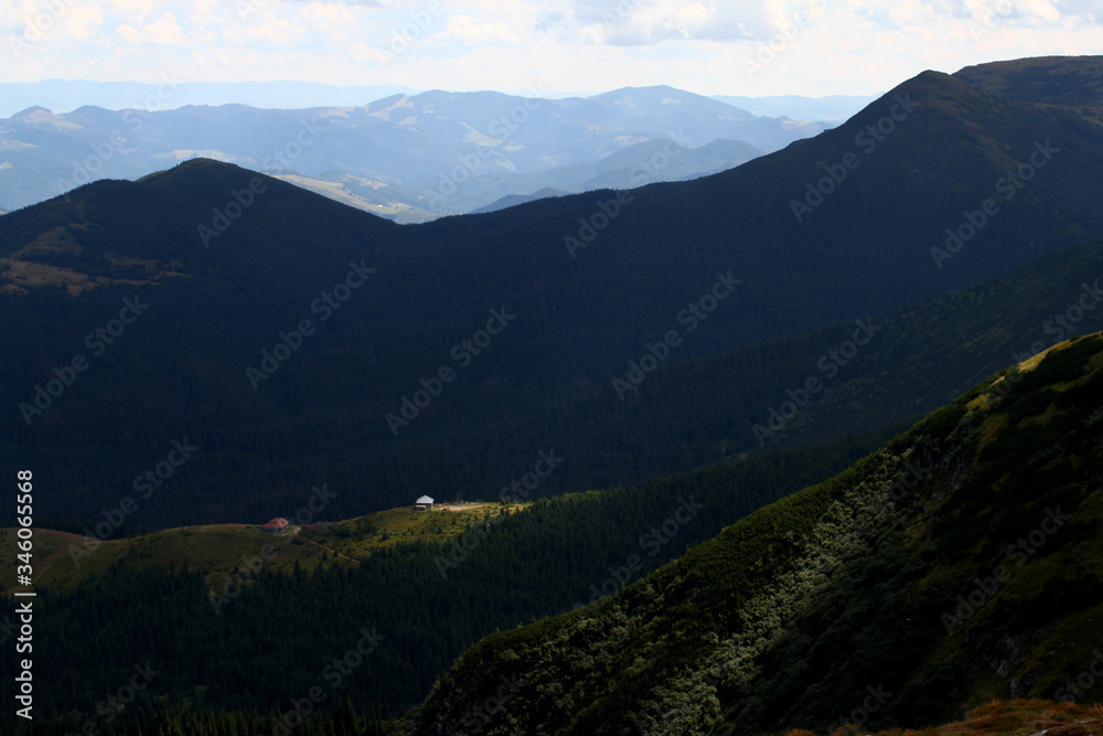 Obraz premium The rays of the sun break through the clouds and illuminate the house standing in a clearing near the edge of the forest among the mountain slopes and peaks. Summer mountain landscape