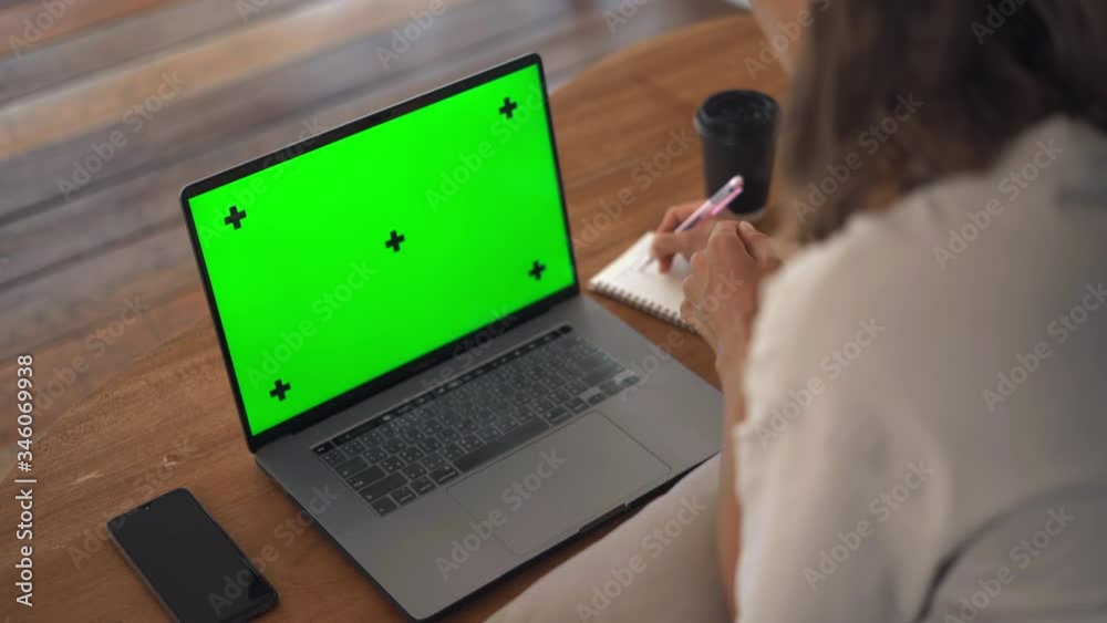 Girl taking notes in notepad from laptop computer Green Mockup Screen. Girl learning online on computer