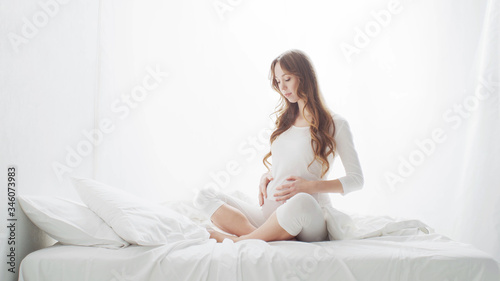 Happy pregnant woman touching her beautiful belly sitting in the bed. Pregnancy, motherhood, and expectation