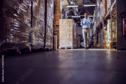 Full length of handsome smiling businessman having phone call with customer while walking in warehouse.