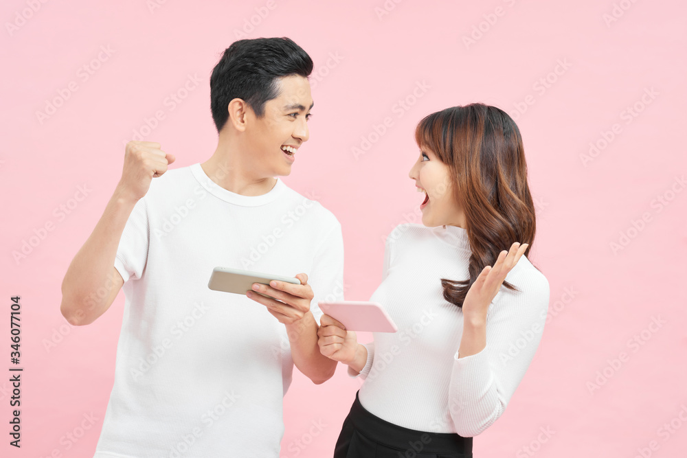 Beautiful couple wearing white t-shirts standing isolated over pink background, playing games on mobile phone. Beautiful couple wearing white t-shirts standing isolated over pink background, playing games on mobile phone.
