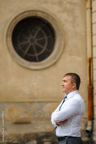 Portrait of a businessman in a white shirt and pants. A man poses on the background of an old building. A man stands sideways and looks in front of him. Hands are gathered in front of the body.