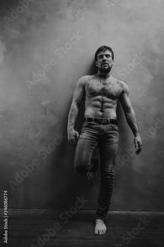 Strong beard man with naked torso posing at the studio wall. Black and white photo.
