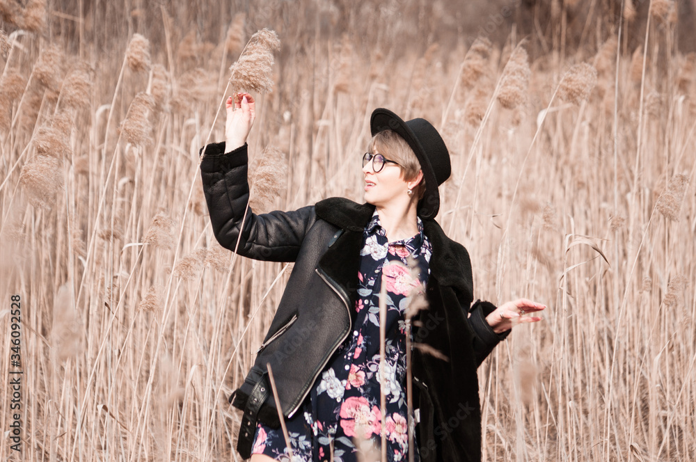 Obraz premium Beautiful happy woman in a hat, glasses and a jacket in the autumn walks in a meadow on a sunny day among dried grass