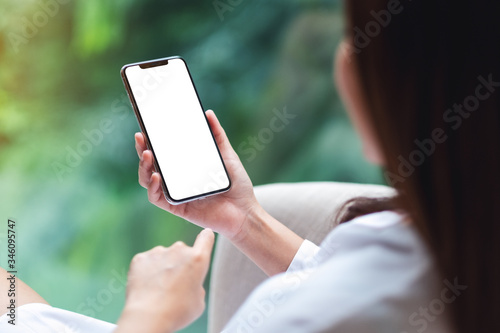 Mockup image of a woman sitting and holding mobile phone with blank white desktop screen , blurred green nature background